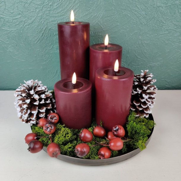 Stemningsfuldt milj�billede af en dekoration p� et rundt fad med bordeaux LED-bloklys arrangeret med gr�nt mos, kogler og r�dbrune b�r.