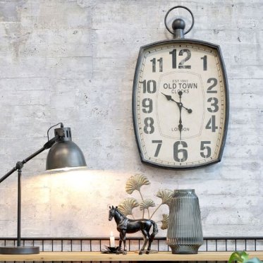 Stort ovalt v�gur i vintage stil med teksten Old Town Clocks London placeret p� en r� betonv�g sammen med en sort bordlampe og dekorative vaser.
