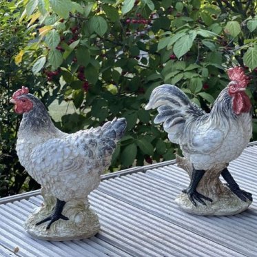 To livagtige havefigurer af en hane og en h�ne i hvide og gr� nuancer med r�de kamme, placeret p� en tr�terrasse med gr�nne buske i baggrunden.