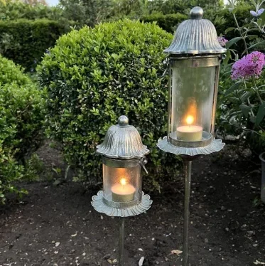 o elegante havelanterner i zink p� spyd med t�ndte fyrfadslys, placeret i et havebed foran en gr�n busk.