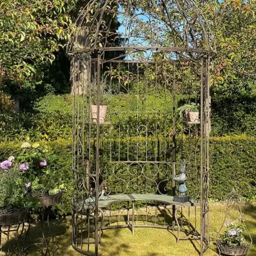 Stor rustik havepavillon i metal med indbygget b�nk, omgivet af gr�nne h�kke, blomsterkrukker og en dekorativ havefigur i en solrig have.