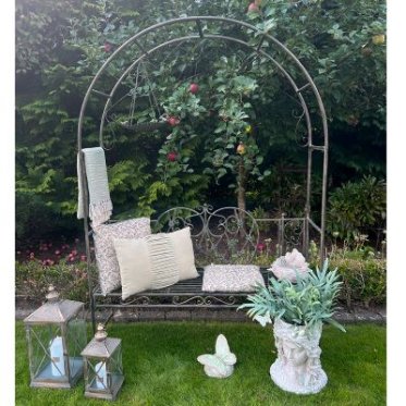 havedekoration-havefigurer-fuglebad-krukker-lanterner-uderum.jpg: Stemningsbillede af et uderum med en haveb�nk under en rosenbue, lanterner p� gr�sset til venstre, en sommerfuglefigur i midten og en krukke formet som et kvindeansigt til h�jre.