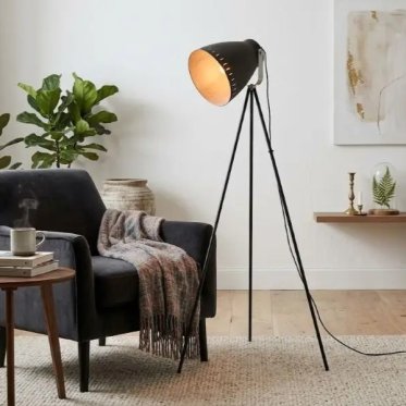 hyggelig-gulvlampe-leitmotiv-tre-ben-nordisk-styling-laesehjoerne.jpg: En t�ndt Leitmotiv trebenet gulvlampe med mat sort finish og guld indersk�rm kaster et varmt, hyggeligt lys ved siden af en m�rkegr� velour-l�nestol med et rustikt strikt�ppe, omgivet 