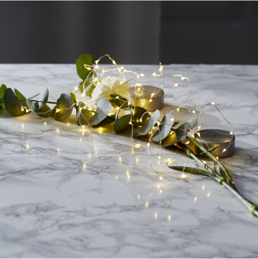 hyggelig-led-lyskaede-eukalyptus-marmor-bord-styling.jpg: En fin tr�d-LED-lysk�de er smukt slynget omkring friske eukalyptusgrene og en hvid nellike p� et lyst marmorbord. Lysene kaster et varmt, hyggeligt sk�r i et roligt og moderne nordisk interi�r.