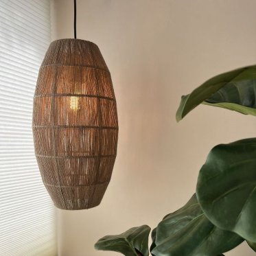 hyggelig-loftlampe-jutesnor-nordisk-landstil-hyggebelysning.jpg: En t�ndt, organisk formet loftlampe v�vet i jutesnor h�nger fra loftet og kaster et varmt, hyggeligt lys. I forgrunden ses de store gr�nne blade fra en stueplante og et hvidt plissegardin, h