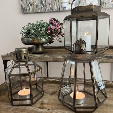 Tre antikke messinglanterner i forskellige st�rrelser med levende lys st�ende p� et rustikt tr�bord ved siden af en dekorativ opsats med planter.