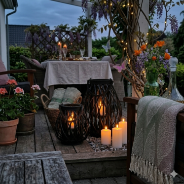 terrassehygge-udeliv-black-bambus-lanterner-led-pillar-plaider-vase-calendula-lys.jpg: Et inspirerende og firkantet stemningsbillede (600x600 px) af en hyggelig terrasseaften med en ops�tning af black bambuslanterner og LED-lys, en gr�n glasvase med Calen