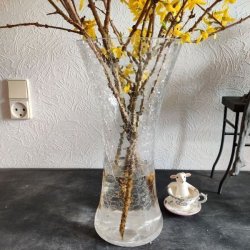 Den store krakelerede glasvase med en buket af gule forsythia-grene er placeret p� et rustikt m�rkt bord. Til h�jre for vasen sidder en lille hvid f�refigur i en m�nstret tekopps-stager, alt sammen foran en m�nstret hvid tapet med et synligt stikkontakt.