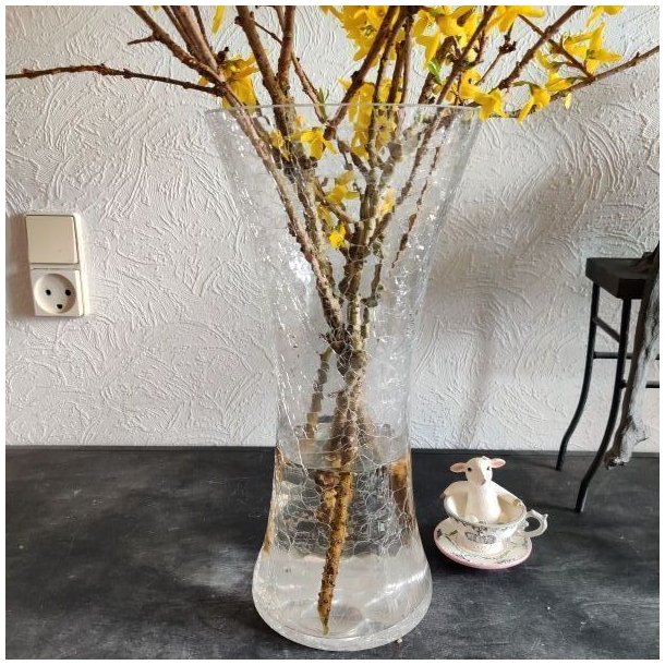 Den store krakelerede glasvase med en buket af gule forsythia-grene er placeret p� et rustikt m�rkt bord. Til h�jre for vasen sidder en lille hvid f�refigur i en m�nstret tekopps-stager, alt sammen foran en m�nstret hvid tapet med et synligt stikkontakt.