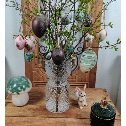 M�rkebrunt metallisk p�ske�g med en fjer, der h�nger p� en for�rsgren i en krystalvase. Vasen st�r p� et tr�bord og er omgivet af andre p�skedekorationer, herunder rosa fjer�g, m�nstrede �g, en kaninfigur og en svampelampe. Salgsproduktet er det brune metalliske �g.
