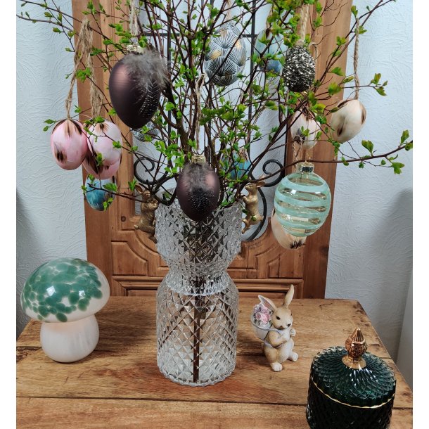 M�rkebrunt metallisk p�ske�g med en fjer, der h�nger p� en for�rsgren i en krystalvase. Vasen st�r p� et tr�bord og er omgivet af andre p�skedekorationer, herunder rosa fjer�g, m�nstrede �g, en kaninfigur og en svampelampe. Salgsproduktet er det brune metalliske �g.