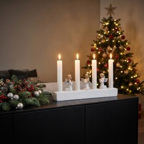Milj�billede af en hvid adventsstage med fire t�ndte lys og tre kn�lende englefigurer, placeret p� en sort sk�nk. I baggrunden ses en juledekoration med gran og kogler samt et juletr� med lys.