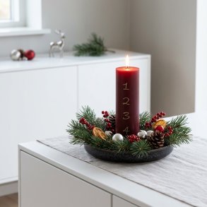 Stemningsfuldt firkantet milj�billede af det t�ndte bordeauxr�de adventslys i en juledekoration med grangrene p� en hvid sk�nk