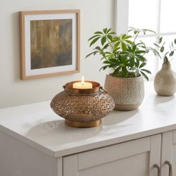 En elegant, antik guld metal hurricane lysestage med et t�ndt stearinlys st�ende p� en hvid sk�nk. Ved siden af er en pottet gr�n plante og et indrammet kunstv�rk. I baggrunden ses et vindue.