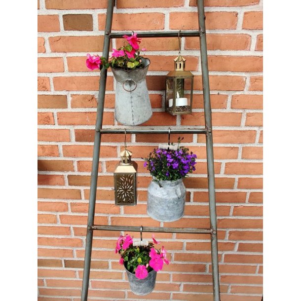 N�rbillede af en dekorativ lanterne i antik messingfinish med et udstanset, orientalsk m�nster p� glasset, her udstyret med et t�ndt LED fyrfadlys. Lanternen h�nger fra en jernstang mod en r�d murstensv�g som en del af en havedekoration p� en stige.