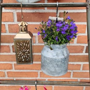 En n�rbillede af en dekorativ lanterne i antik messing finish, der h�nger fra en jernstang mod en r�d murstensv�g. Lanternen har glaspaneler med et fint udstanset, orientalsk m�nster p� metall�gen. Til h�jre for den h�nger en zink m�lkejunge med bl� lobelia blomster.