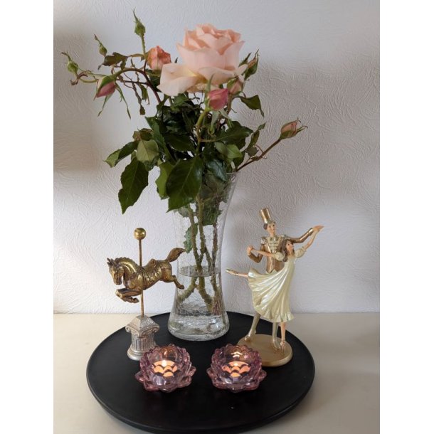 Et kvadratisk milj�billede i 1:1 format af en elegant dansende balletfigur i guld og creme. Figuren er placeret p� en sort rund bakke ved siden af en vase med ferskenfarvede roser, en gylden karruselhest og to t�ndte lys i lyser�de lotusformede glasholdere mod en hvid v�g.