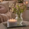 Et firkantet milj�billede, der viser det store fransk gr� bloklys med 3 t�ndte v�ger placeret p� en glasplade. Ved siden af lyset st�r en mindre vase med blomster, og i baggrunden anes en sofa med lyser�de hynder i et hyggeligt stuemilj�.
