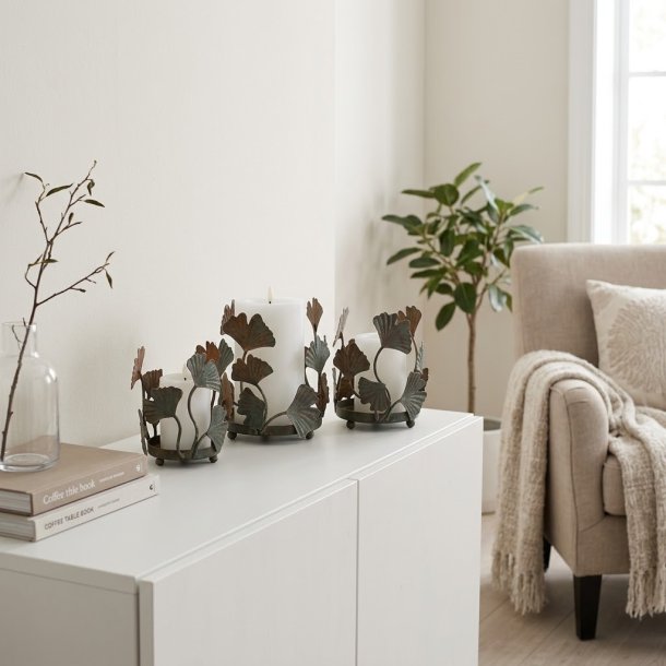 Stemningsfuldt firkantet milj�billede af de tre bloklysestager m/ bladdekoration i antique messing placeret p� en hvid sk�nk i stuen med t�ndte hvide LED-bloklys.