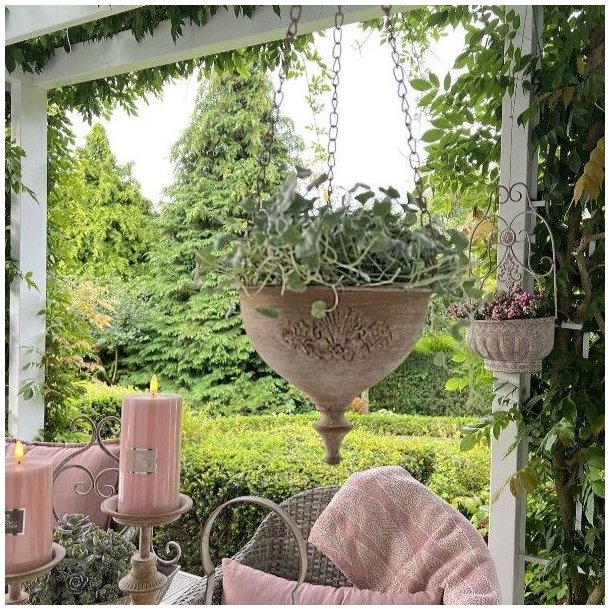 En rustik blomsterpotte i antik sandfarvet jern h�nger dekorativt i en gr�n have. Potten er monteret med en metalk�de og indeholder en lille gr�n plante.