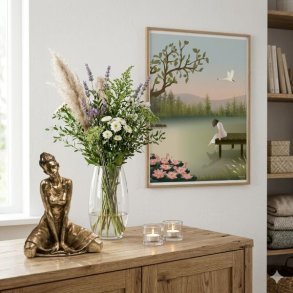 Et hyggeligt 1:1 milj�billede, der viser en curated tr�sk�nk. P� venstre side st�r en detaljeret bronze-skulptur af en siddende ballerina. Ved siden af hende st�r en elegant glaspokalvase fyldt med friske pampasgr�s og blomster. To sm� glasfyrfadslysholdere med t�ndte stearinlys fuldender opstillingen. Centreret p� v�ggen over sk�nken h�nger en indrammet Vissevasse-plakat med motivet af en kvinde p� en bro ved en s�, hvilket binder hele rummet sammen under bl�dt, naturligt dagslys.