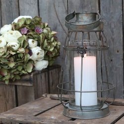 Et stemningsfuldt milj�billede i 1:1 format af en Factory lanterne i antique zink fra Chic Antique. Lanternen er t�ndt med et hvidt bloklys og st�r p� et rustikt tr�bord ved siden af en frodig buket blomster.