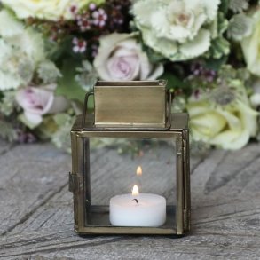 Et hyggeligt indend�rs milj�billede, hvor den lille messinglanterne st�r t�ndt med et fyrfadslys p� et rustikt tr�bord ved siden af en lille blomsterdekoration.