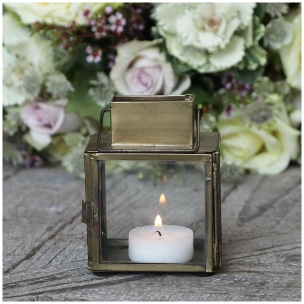Et hyggeligt indend�rs milj�billede, hvor den lille messinglanterne st�r t�ndt med et fyrfadslys p� et rustikt tr�bord ved siden af en lille blomsterdekoration.