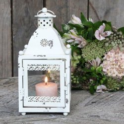 Stemningsbillede af den hvide lanterne placeret p� et rustikt tr�bord med et t�ndt lys indeni ved siden af en smuk blomsterbuket.