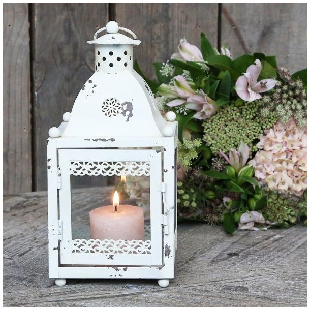 Stemningsbillede af den hvide lanterne placeret p� et rustikt tr�bord med et t�ndt lys indeni ved siden af en smuk blomsterbuket.