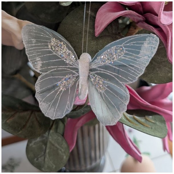 Gr�bl� pyntesommerfugl med glimmer sat fast p� m�rkegr�nne blade og lyser�de blomster i en vase.