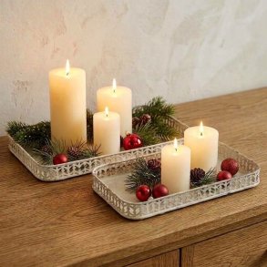 Firkantet milj�billede af to rektangul�re dekorationsfade i antik creme jern med udstanset kant og patina, placeret p� en tr�sk�nk og pyntet med t�ndte hvide bloklys og grangrene uden t�ppe i baggrunden.