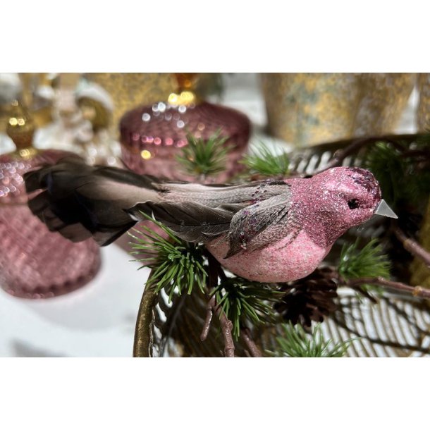 Rosabrun dekorationsfugl med glimmer og lange fjer placeret p� en grangren med dekorative rosa glasb�tter i baggrunden.