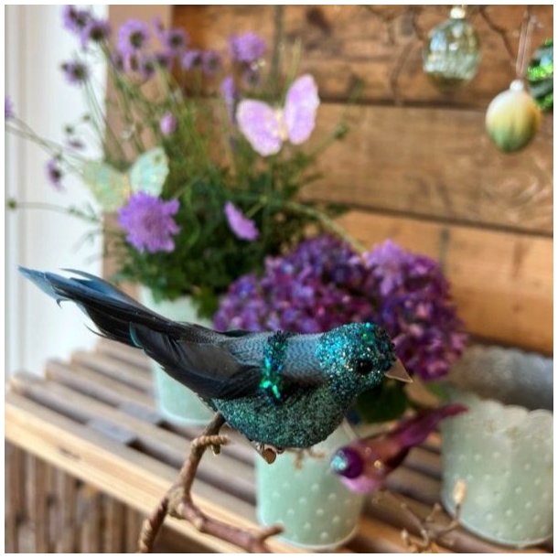Flot dekorativ fugl i mint og bl�gr�nne nuancer med glimmer placeret p� en gren i en hyggelig indretning med lilla blomster.