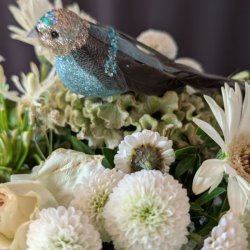 N�rbillede af turkisbl� dekorativ fugl med glitrende s�lvhoved og turkist pailletb�nd, placeret i en t�t hvid blomsterbuket.