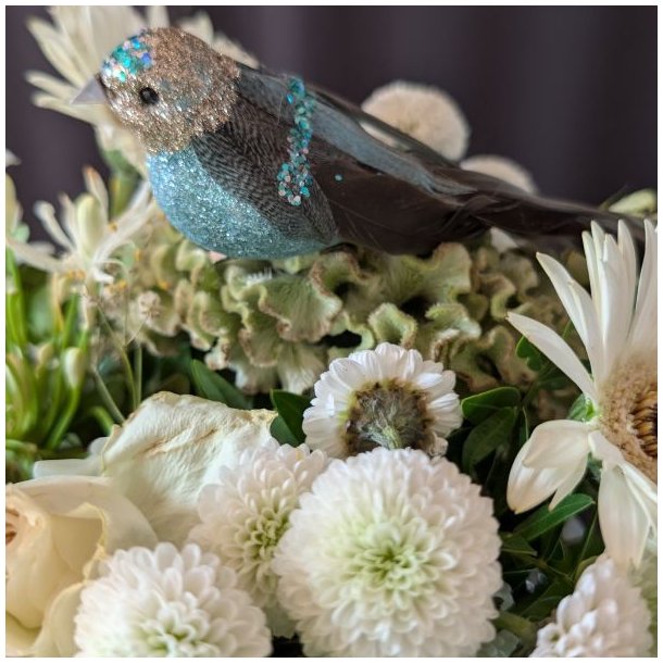 N�rbillede af turkisbl� dekorativ fugl med glitrende s�lvhoved og turkist pailletb�nd, placeret i en t�t hvid blomsterbuket.