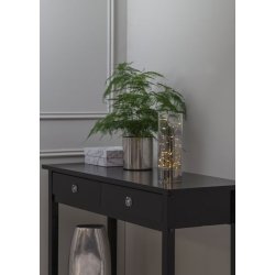 Et milj�billede af en lysende glasvase med timer-funktion placeret p� et sort konsolbord ved siden af en gr�n plante og dekorative �sker i en moderne stue.