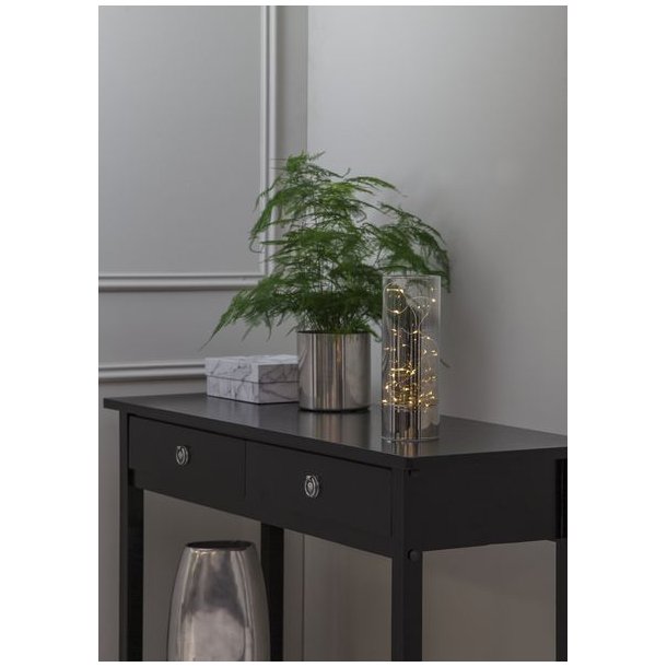 Et milj�billede af en lysende glasvase med timer-funktion placeret p� et sort konsolbord ved siden af en gr�n plante og dekorative �sker i en moderne stue.