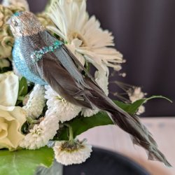 Profilbillede af en 15 cm lang turkisbl� pyntefugl med m�rk fjerhale og masser af glimmer i en hvid blomsteropsats.