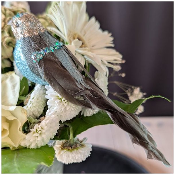 Profilbillede af en 15 cm lang turkisbl� pyntefugl med m�rk fjerhale og masser af glimmer i en hvid blomsteropsats.