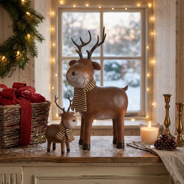 Et hyggeligt julemilj�billede i en vindueskarm med de to brune rensdyr-figurer Romeo fra Det Gamle Apotek st�ende sammen. Det store rensdyr p� 31 cm og det lille rensdyr p� 17 cm er placeret p� et rustikt tr�bord med kunstig sne, et stribet t�rkl�de, grankogler og et t�ndt fyrfadslys, med et snekl�dt sprossevindue i baggrunden.