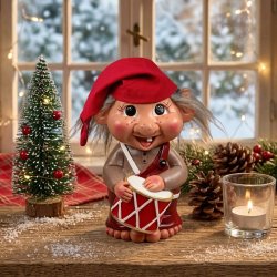 Et hyggeligt julemilj�billede i en vindueskarm med troldedrengen Jeppe p� 22 cm fra Det Gamle Apotek i centrum. Trolden har en r�d nissehue, vildt gr�t h�r og en r�d-hvid tromme. Den st�r p� et rustikt tr�bord omgivet af et lille juletr� med lys, grankogler og en lysestage fra Det Gamle Apotek med et t�ndt fyrfadslys, med et snekl�dt sprossevindue i baggrunden