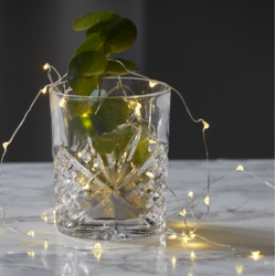 Et krystalklart whiskyglas med indviklet slibning st�r p� en hvid marmorbordplade mod en m�rkegr� baggrund. I glasset er der friske gr�nne eukalyptusblade. En delikat lysk�de med varme, gyldne LED-lys er v�vet ind imellem bladene, viklet rundt om glasset og ligger dekorativt p� bordet omkring det. Lysk�dens lys reflekteres smukt i det sk�rne glas.