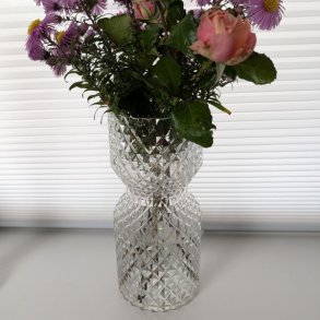 diamantslebet-glas-vase Glasvase med et flot diamantslebet m�nster, formet som et timeglas. Vasen indeholder en buket med lyser�de blomster og st�r p� en lys overflade foran en hvid persienne.