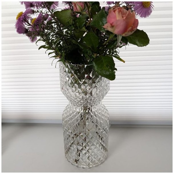 diamantslebet-glas-vase Glasvase med et flot diamantslebet m�nster, formet som et timeglas. Vasen indeholder en buket med lyser�de blomster og st�r p� en lys overflade foran en hvid persienne.