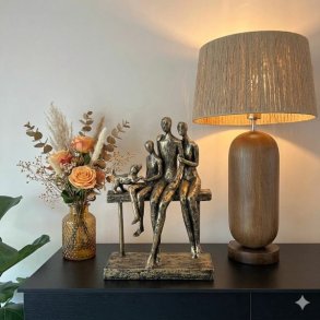 Et stemningsfuldt milj�billede p� en sort sk�nk. Centralt ses en patineret bronze-skulptur af en familie p� tre personer og en hund, der sidder sammen p� en b�nk. Til h�jre for figuren st�r en oplyst bordlampe med en cylinderformet valn�ddetr�-base og en t�tv�vet raffia-sk�rm. Til venstre er en tekstureret ravfarvet glasvase med en buket af t�rret pampasgr�s og ferskenfarvede roser. Baggrunden er en lys v�g, og en plante ses i forgrunden. Belysningen er varm og hyggelig.