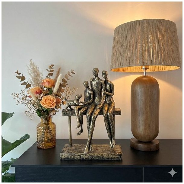 Et stemningsfuldt milj�billede p� en sort sk�nk. Centralt ses en patineret bronze-skulptur af en familie p� tre personer og en hund, der sidder sammen p� en b�nk. Til h�jre for figuren st�r en oplyst bordlampe med en cylinderformet valn�ddetr�-base og en t�tv�vet raffia-sk�rm. Til venstre er en tekstureret ravfarvet glasvase med en buket af t�rret pampasgr�s og ferskenfarvede roser. Baggrunden er en lys v�g, og en plante ses i forgrunden. Belysningen er varm og hyggelig.