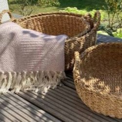 Et indbydende, kvadratisk milj�billede taget i fuldt dagslys p� en terrasses tr�gulv. To af de flettede s�gr�skurve med h�ndtag fra La Vida st�r her. Den st�rste kurv (L; D45 cm) er fyldt med et hyggeligt, lyser�dt t�ppe med frynser. Ved siden af st�r den mellemst�rste kurv (M; D40 cm), tom og klar til brug. I baggrunden ses en frodig, gr�n have og terrassestole, der skaber en afslappet og funktionel atmosf�re. S�gr�ssets naturlige farve og struktur st�r skarpt frem.