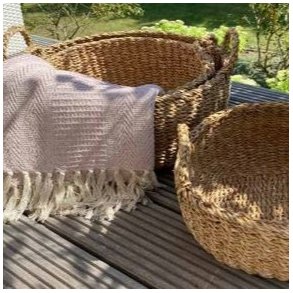 Et indbydende, kvadratisk milj�billede taget i fuldt dagslys p� en terrasses tr�gulv. To af de flettede s�gr�skurve med h�ndtag fra La Vida st�r her. Den st�rste kurv (L; D45 cm) er fyldt med et hyggeligt, lyser�dt t�ppe med frynser. Ved siden af st�r den mellemst�rste kurv (M; D40 cm), tom og klar til brug. I baggrunden ses en frodig, gr�n have og terrassestole, der skaber en afslappet og funktionel atmosf�re. S�gr�ssets naturlige farve og struktur st�r skarpt frem.