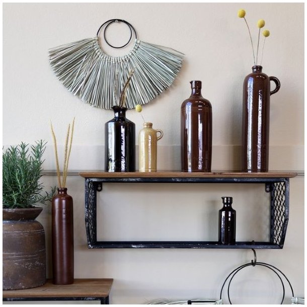 En dekorativ v�gopstilling med forskellige brune keramikvaser og flasker p� en sort metalhylde. Den store flaske med hank st�r til h�jre med tre gule blomster i.